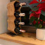 An oak wood tabletop wine rack holding 4 bottles of red wine. The wine rack is placed next to a window with a red plant.